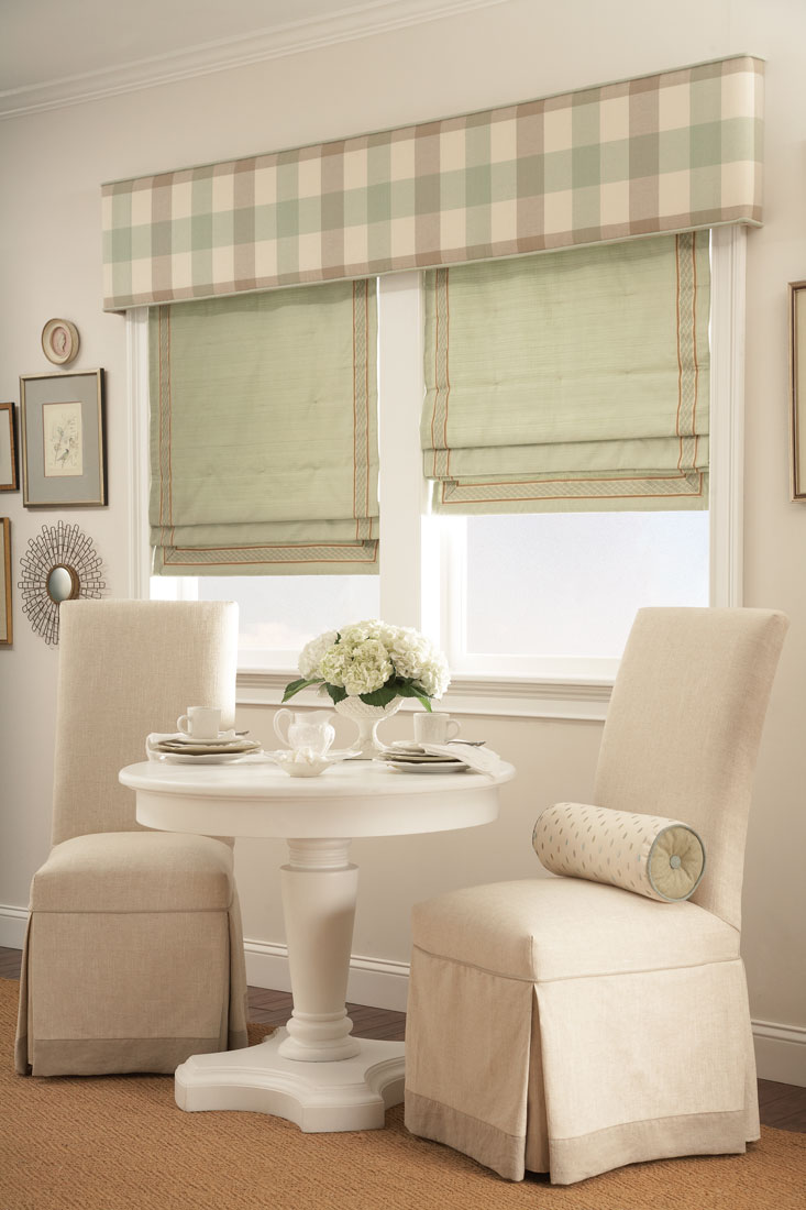Two windows with light green Interior Masterpieces® Fabric Shades & accenting Fabric Wrapped Cornice spanning both of them behind a couple tan chairs and white table