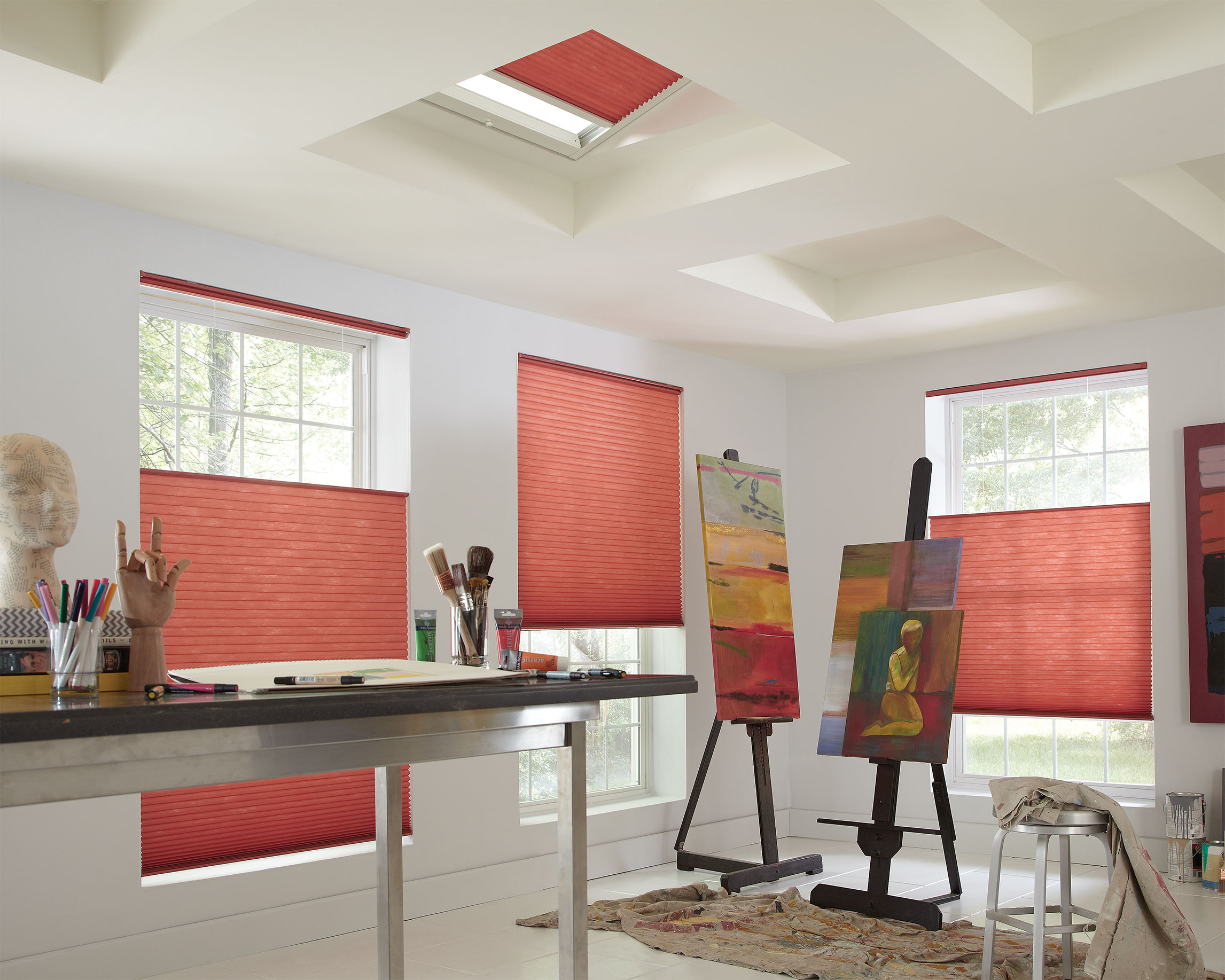 Bright rose colored cellular shades hang in three large windows and the skylights of an artist's studio featuring bright paintings and tarps.