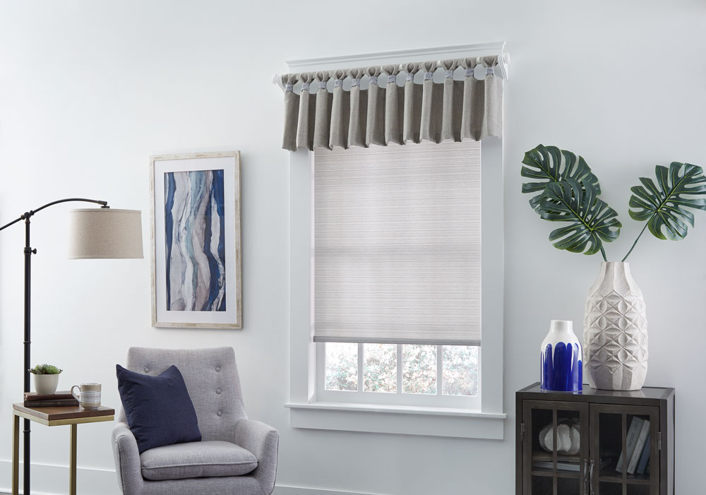 White Genesis® roman shade with Interior Masterpieces valance hanging on a white custom rod with finials in a room with white walls and a blue painting and white chair with a blue Interior Masterpieces® accenting pillow