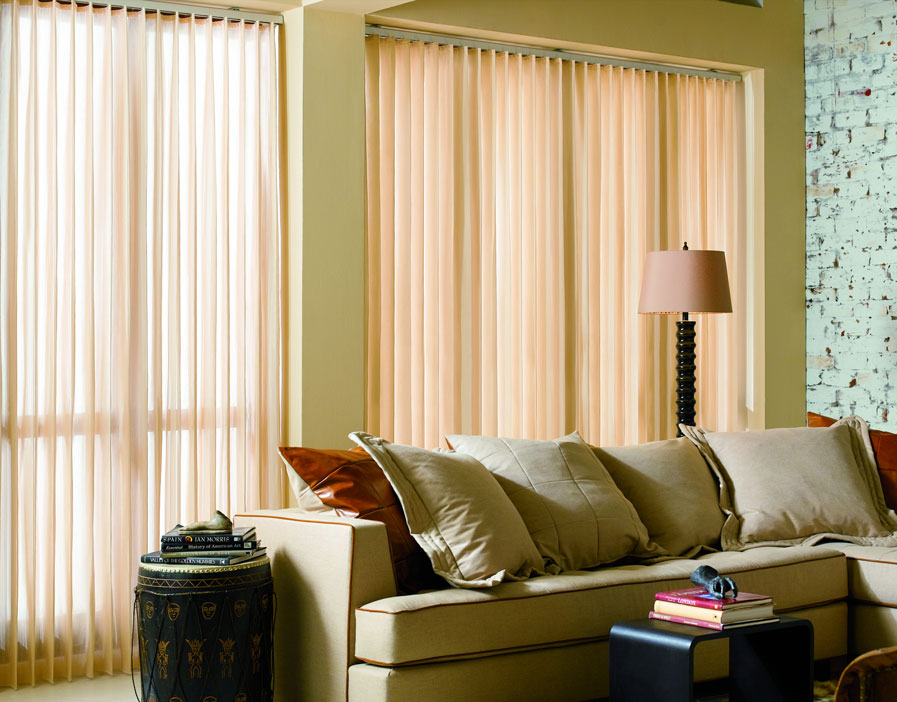 Two large Sheer Visions® Vertical Blinds hanging in a window with a couch and lamp in front