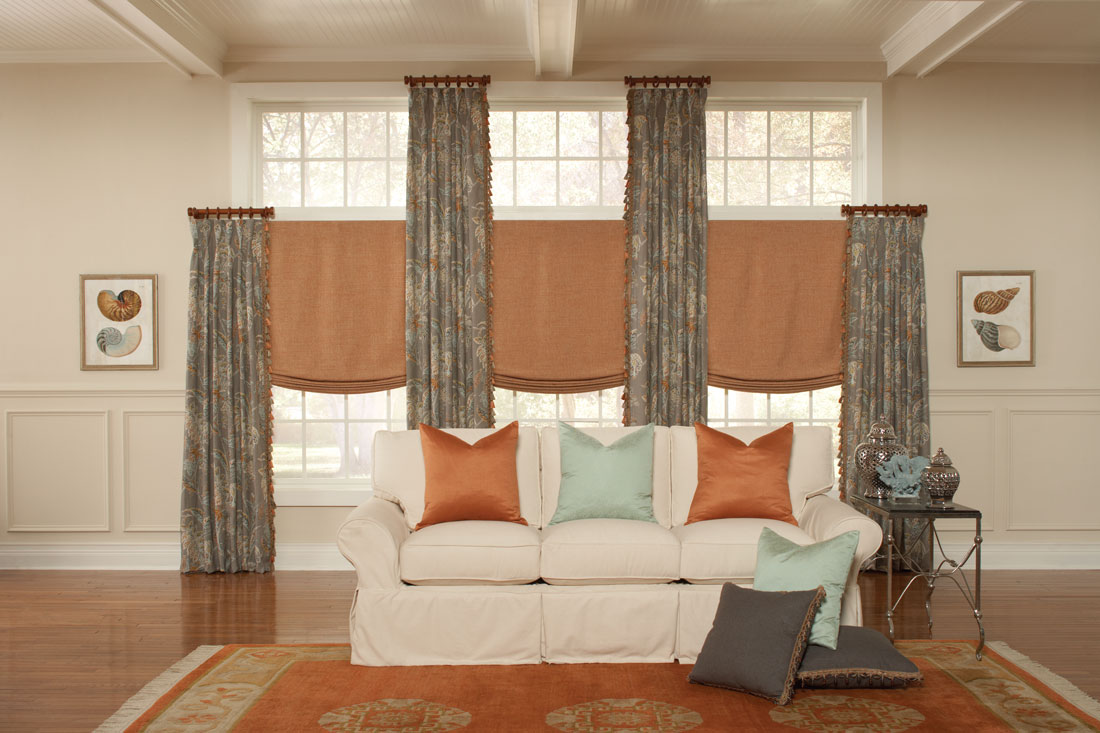 dark orange Interior Masterpieces® fabric shades with gran floral patterned draperies hanging on dark brown custom rods with rings and finials and accenting custom pillows on a couch