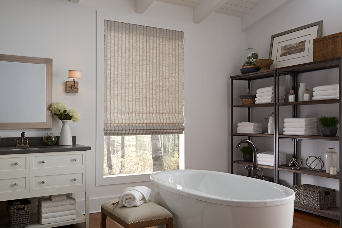 a tan Genesis® Hobbled Roman Shade in a bathroom behind a white stand alone tub next to shelves with towels