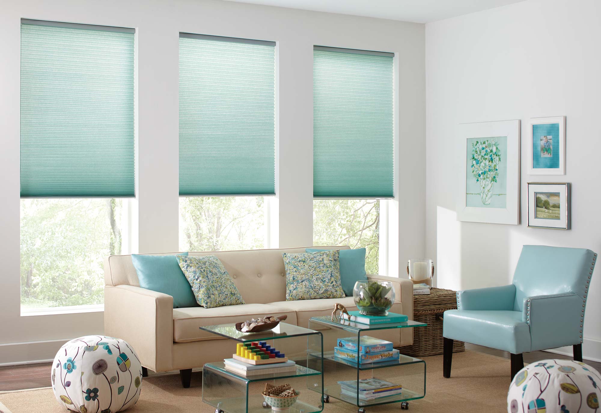 Three aqua colored Parasol® Cellular Shades hang in the windows of a bright family room with custom blue and patterned pillows on a modern cream couch.