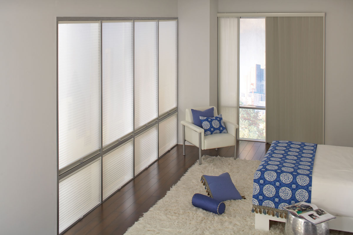 Large picture windows on one side of the room with Parasol® Duo-Lucent Cellular shades in them and a Parasol® Glissade panel system on the other in a light tan material with a bed and chair that have custom bedding in a blue material