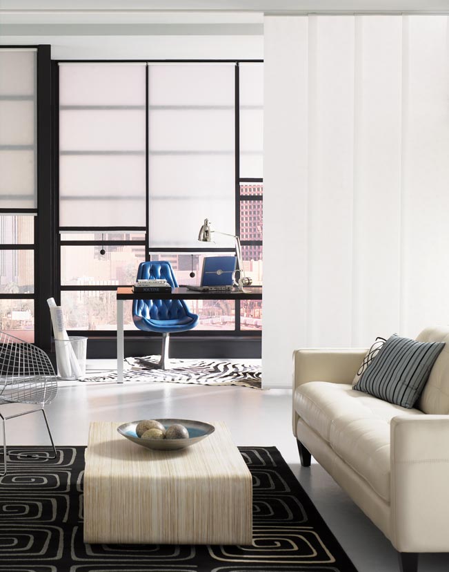 several white Genesis® Roller Shades with black Banding in an office behind a desk and a Panel Track acting as a room divider  with white panels behind a white couch and dark rug