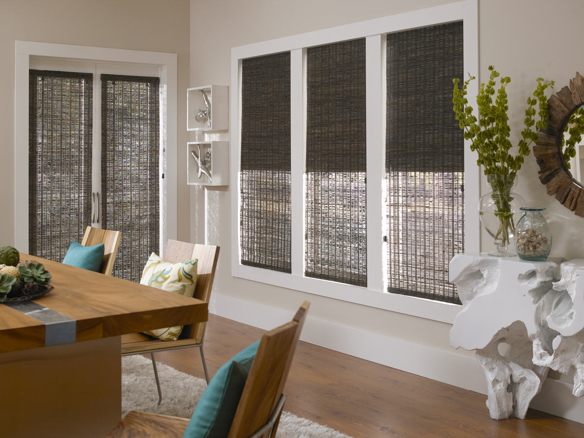 Several dark Manh Truc® Woven Wood Shades in a dining area with a brown table and chairs that have blue and green floral patterned custom pillows on them