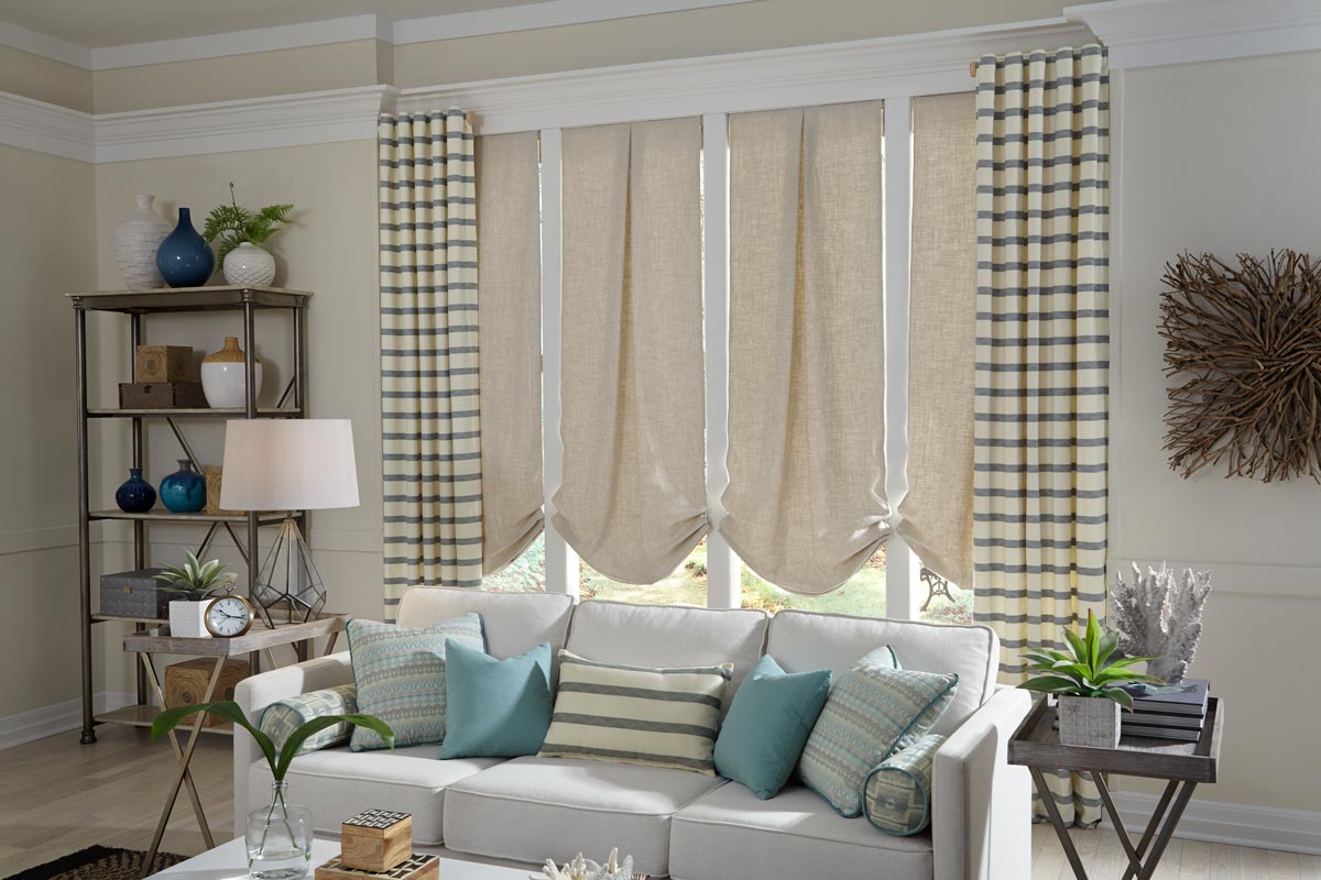 4 tan Interior Masterpieces® Roman Shades with pleats and back tab draperies hanging on either side behind a couch that has several different light green and blue custom pillows