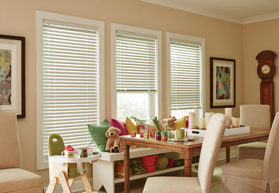 3 white Classic Collection® Aluminum Blinds in a dining room with children's toys and a high chair with colorful Interior Masterpeices® Custom Pillows on a bench in front