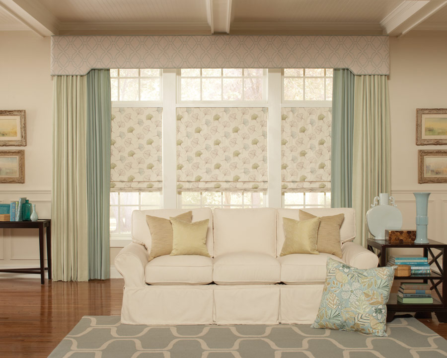 Interior Masterpieces® cornice spanning a large picture window with matching draperies and fabric shades underneath with a couch that has custom pillows in front