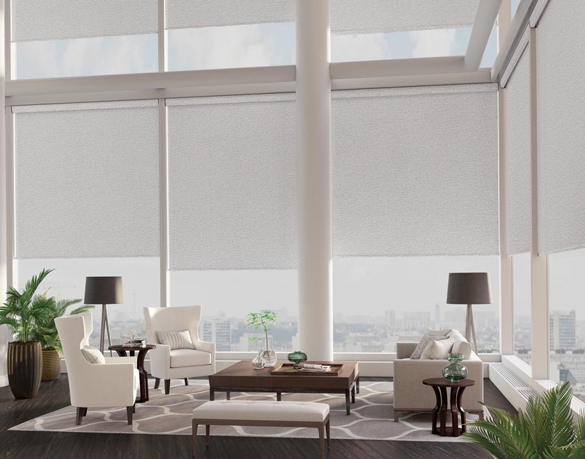 Several tan colored Genesis® Motorized Roller Shades in a high rise corner dining area with white chairs and a brown table in the foreground