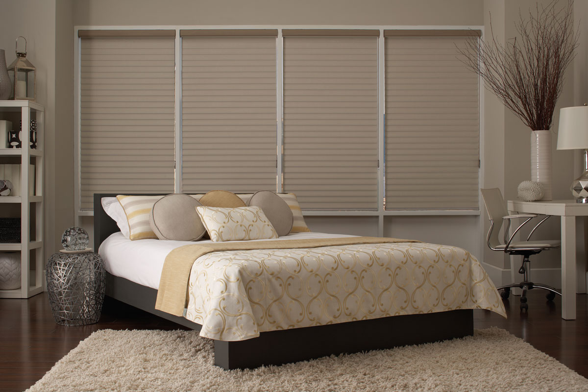 Several tan Tenera® sheer shades in the closed position in a tan bedroom with a bed that has custom Interior Masterpieces® pillows and blankets on it