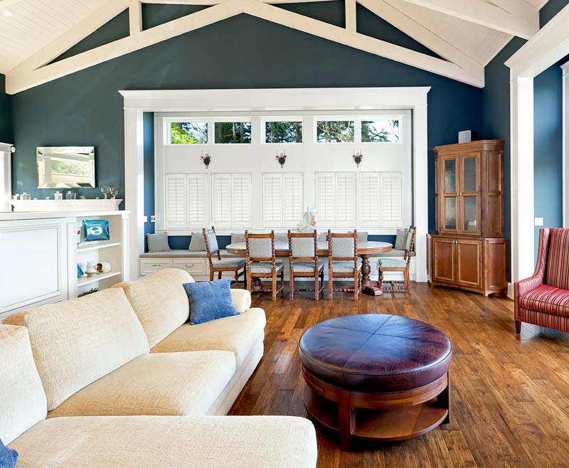 several white Parke® Shutters against a blue wall in a dining area with brown chairs and a table nearby and a cream colored couch in the foreground