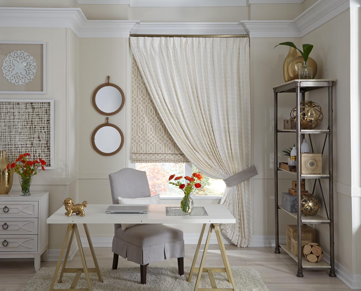 A light colored Pleated Interior Masterpieces® drapery hanging in front of a slightly darker geometric patterned flat roman shade in a room with a desk and laptop with a chair and book shelf full of  decorative pieces