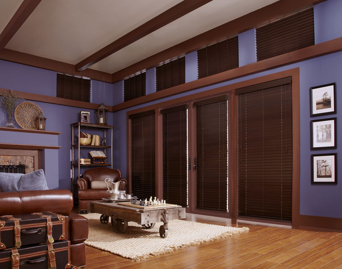 several dark brown Heartland Woods® Wood Blinds with Valances against a purple wall with brown lather furniture in the room