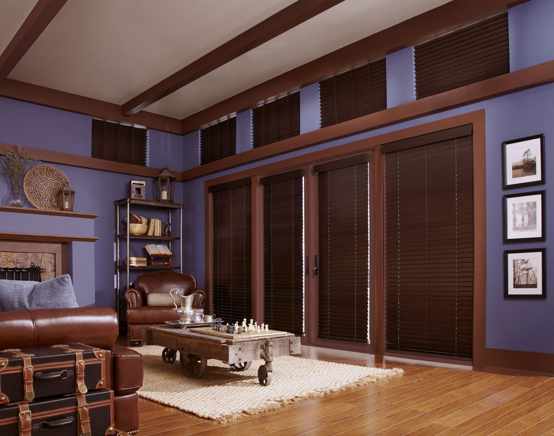 several dark brown Heartland Woods® Wood Blinds with Valances against a purple wall with brown lather furniture in the room