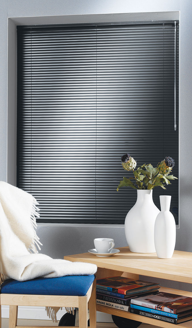 Dark Classic Collections® Aluminum Blinds in a room with light gray walls with a chair and table in the foreground