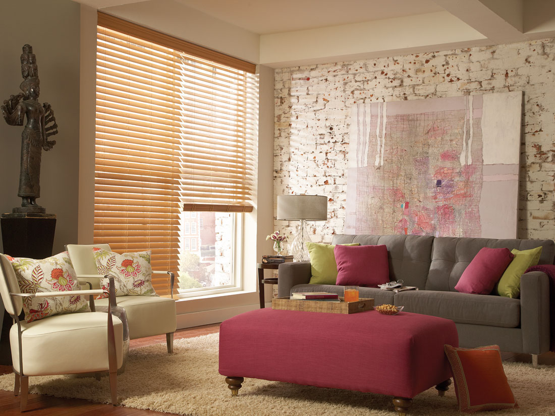 light Brown Fidelis® faux wood blinds in a room with a gray couch and maroon ottoman with green and maroon custom pillows on it next to white chairs with floral printed custom pillows