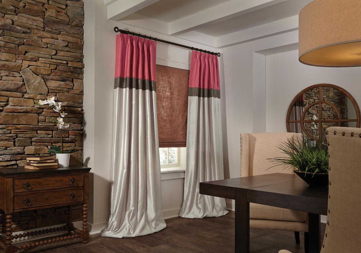 Long flowing light red and tan Interior Masterpieces® draperies pooling on the floor in front of a red flat roman shade