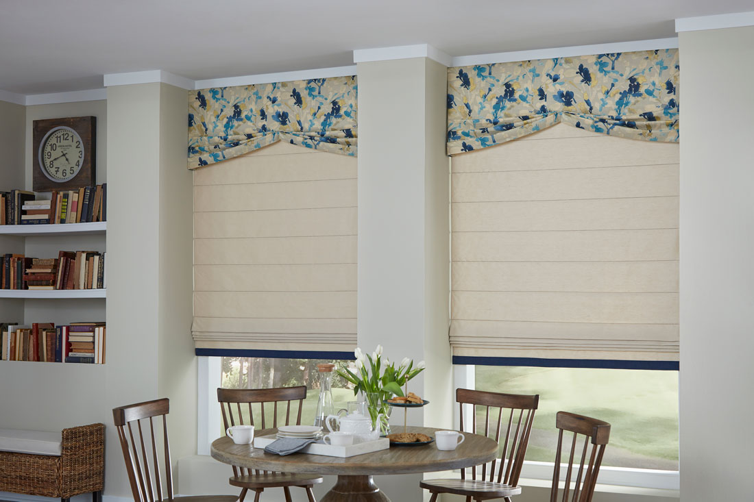 Two tan Interior Masterpieces® fabric shades with custom fabric valances that have a blue and yellow floral pattern behind brown table and chairs