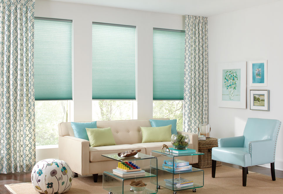 Three Parasol® Cellular shades in a light blue color with accenting Custom Interior Masterpieces® draperies hanging on each end in front of a tan couch with matching pillows and a blue matching chair