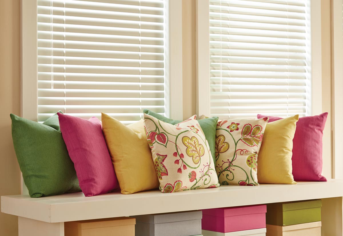 Several brightly colored Interior Masterpieces® Custom Pillows in front of white Classic Collection® Aluminum Blinds