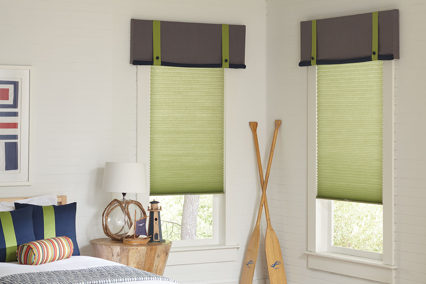 Light, grassy green cellular shades hang in the two corner windows of a child's bedroom featuring nautical accessories, custom matching valances in gray and pillows in navy with green accents.