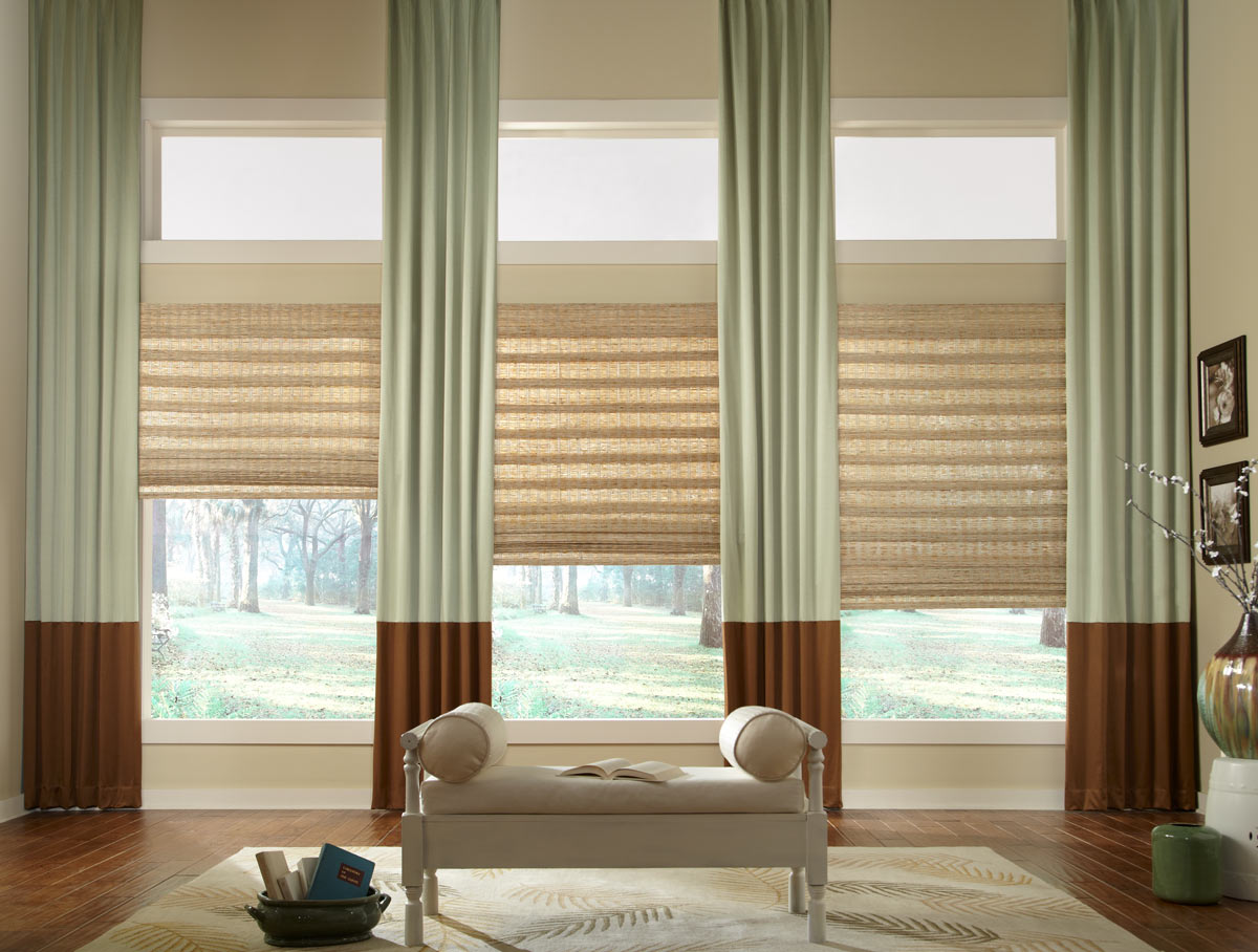 several light brown Manh Truc® Woven Wood Shades with light green and dark brown Interior Masterpieces® Draperies hanging between with a light brown chair in the foreground