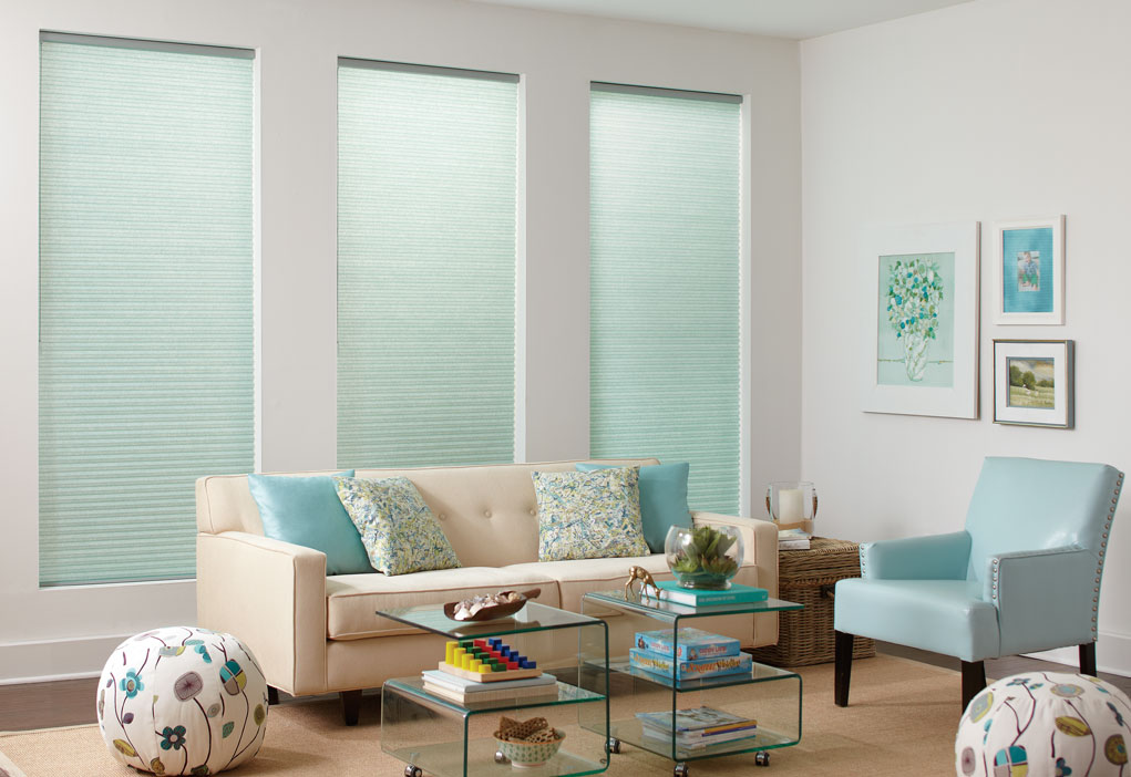 Three Parasol® Cellular shades in a light blue color in front of a tan couch with matching pillows and a blue matching chair