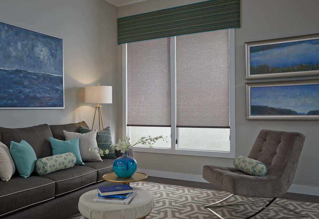 Two large gray Genesis® Roller Shades with a green and yellow striped Interior Masterpieces® Fabric Cornice spanning both shades and a dark gray couch with accenting blue, gray and green Custom Pillows