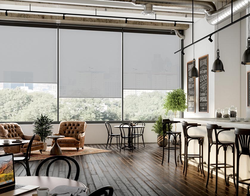 Several dark Genesis® Motorized Roller Shades in a restaurant with stools and a seating area with brown chairs and tables