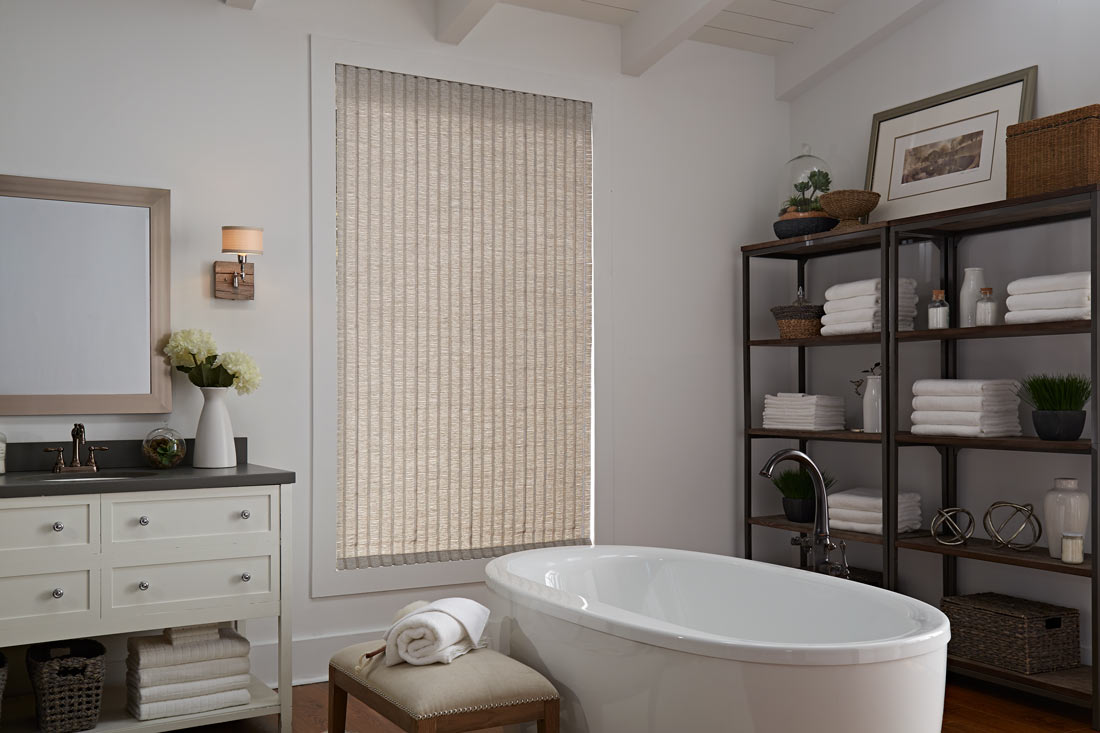 a tan Genesis® Hobbled Roman Shade in a bathroom behind a white stand alone tub next to shelves with towels