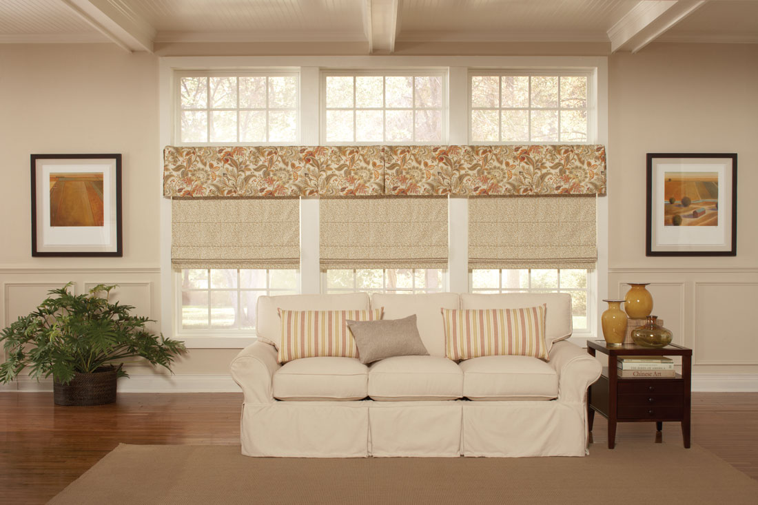 three cream colored Interior Masterpieces® fabric shades with a floral patterened cornice spanning the width of all three above behind a white couch tha thas custom accent pillows