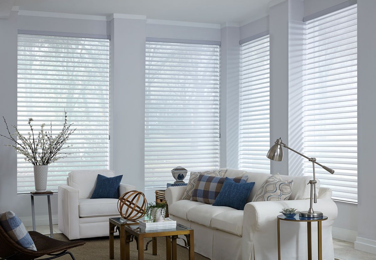 Several white Tenera® Sheer Shades in the open position in a room with white walls and a white couch with a white chair that have blue and blue plaid Interior Masterpieces® custom pillows