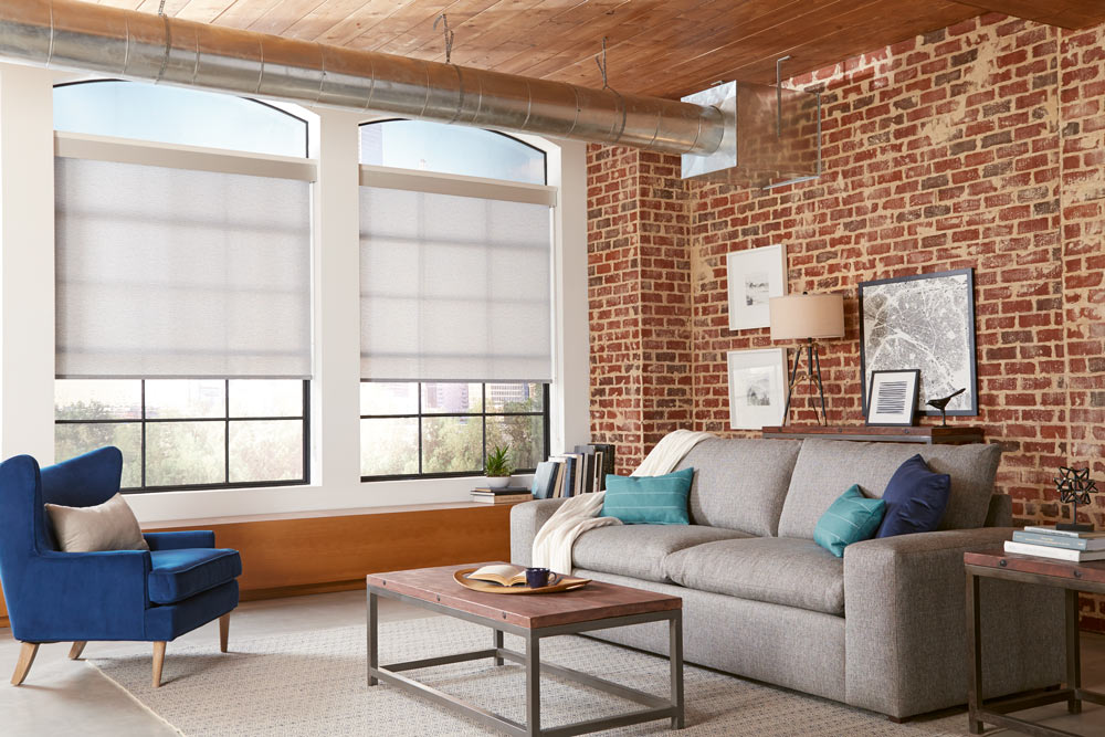 two tan Genesis® Roller Shades against brick walls with a gray couch that has blue and teal Interior Masterpieces® Custom Pillows