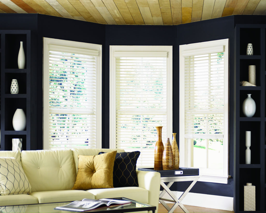 3 white Heartland Woods® Wood Blinds in a bay window against blue walls with a white couch that has Interior Masterpieces® custom pillows on it