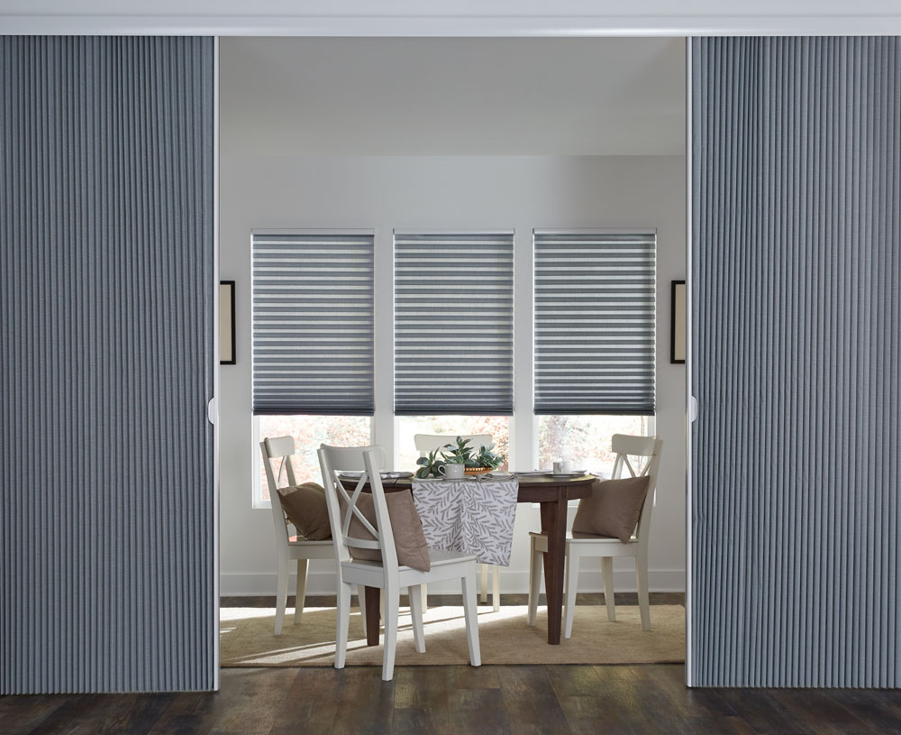 three blue Parasol® Cellular Shades against a white wall with a  Glissade Panel system in front separating a dining area with white chairs a brown table and Interior Masterpieces® custom pillows