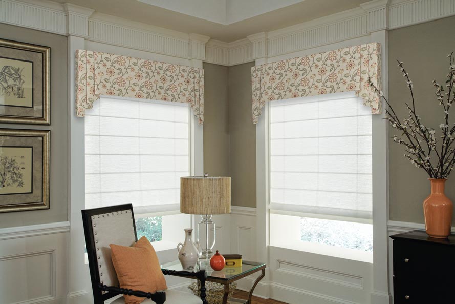 Two white Genesis® Roman Shades with tan floral patterened Interior Masterpieces Valances and a chair that has a peach colored Custom Pillow