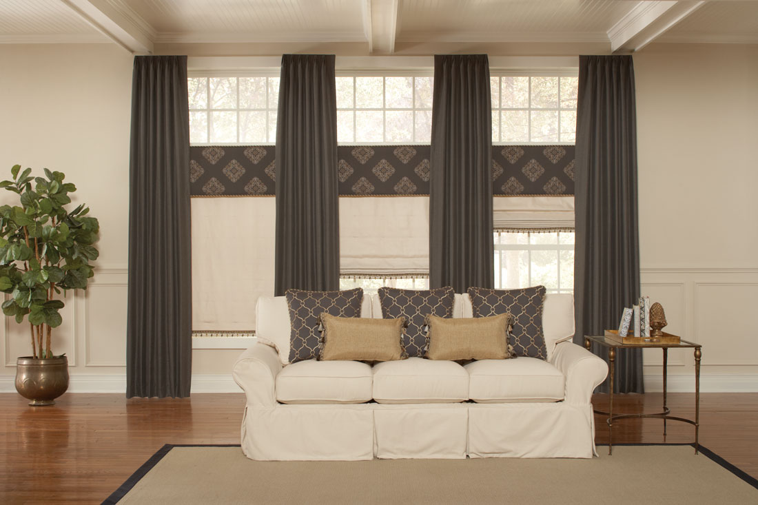 three tan Interior Masterpieces® fabric shades with Embellishment trim tassles and dark brown fabric wrapped cornices above with long flowing dark brown draperies between each behind a white couch with custom accent pillows