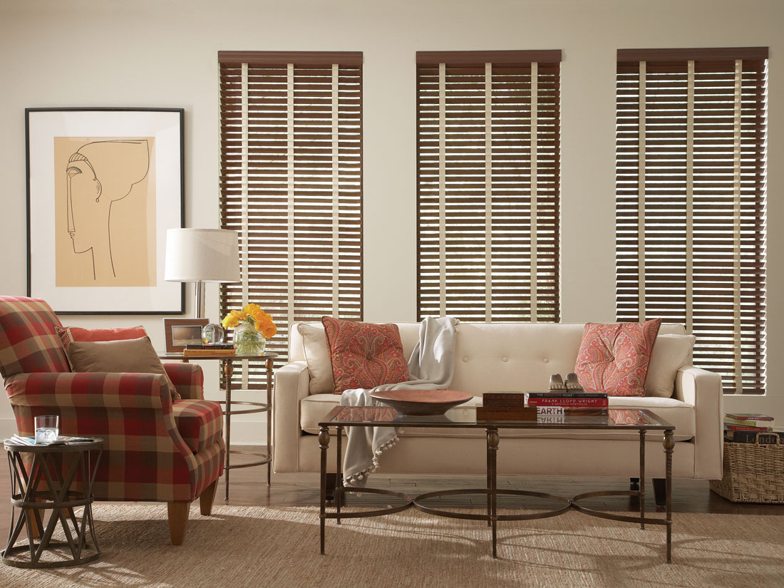 three dark brown Fidelis® Faux Wood Blinds with light colored Banding in an room with a white couch and red plaid chair with Interior Masterpieces® pillows on them
