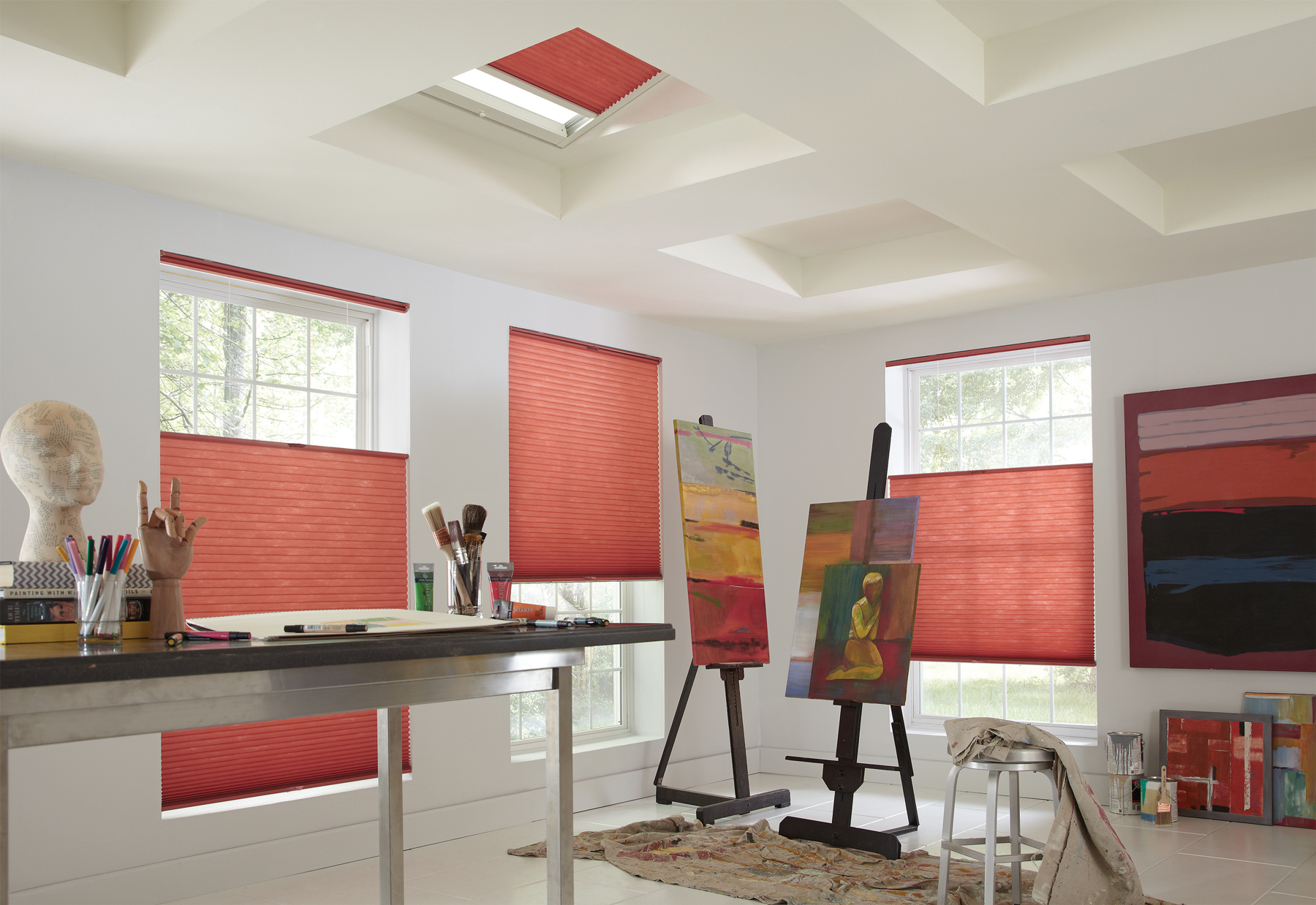 Three red Parasol® Top Down-Bottom Up shades in windows with a Parasol SkyLight shade in the ceiling in a studio room with painting supplies and paintings nearby