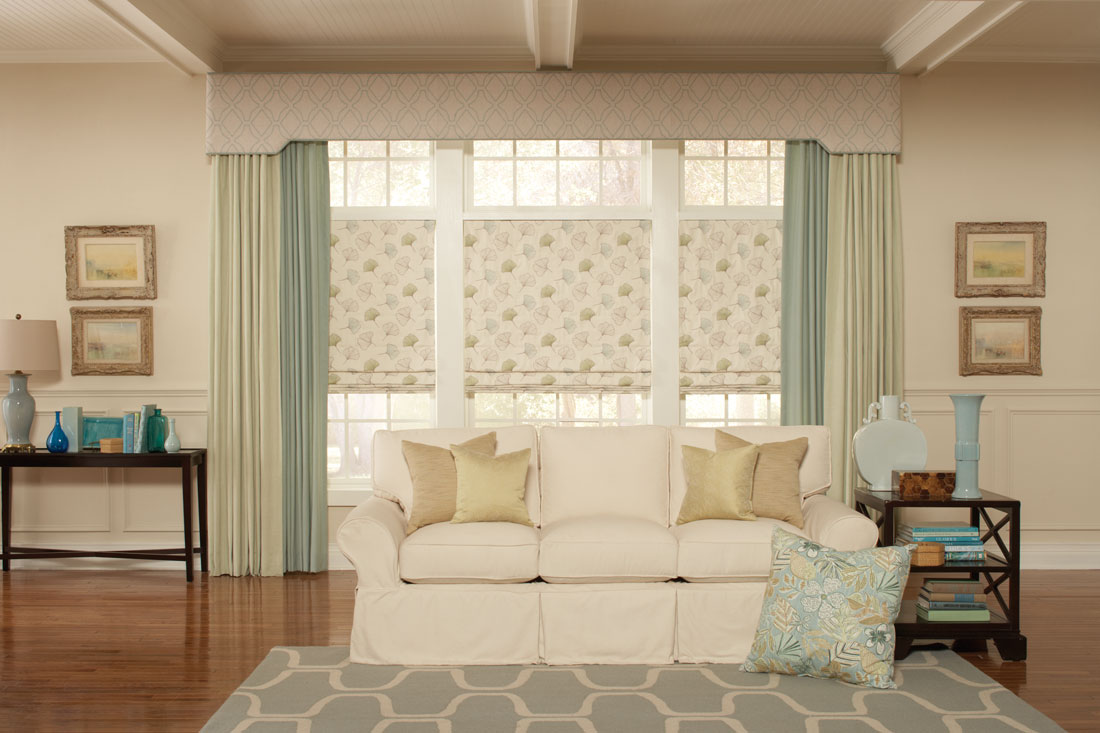 three tan floral patterned Interior Masterpieces® fabric shades with a large fabric wrapped cornice spanning the windows with light and dark green draperies hanging at each end behind a white couch with custom pillows