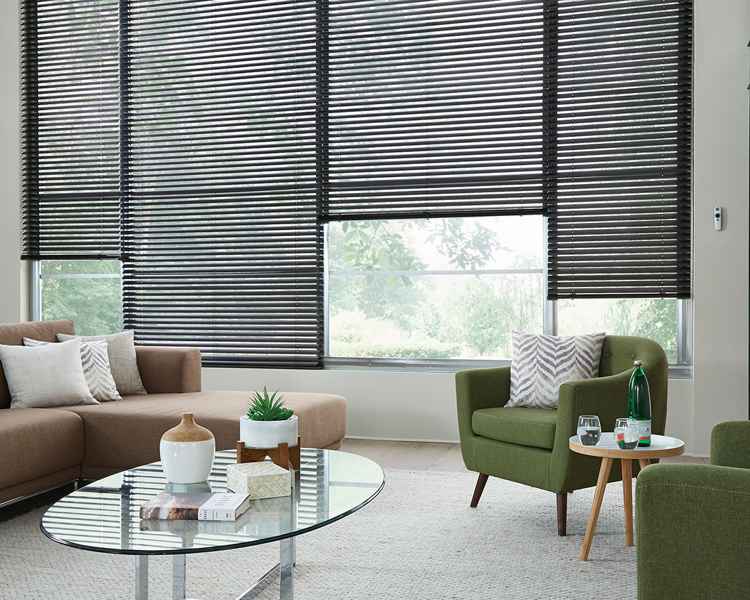 Dark Classic Collection® Motorized Aluminum Blinds in a room with a brown couch, green chair and Interior Masterpieces® pillows