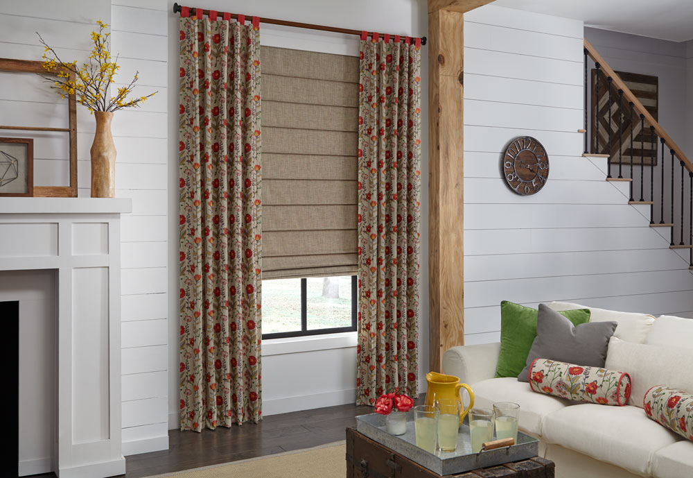 red and tan floral patterned Interior Masterpieces® draperies hang on a dark wooden custom rod and finials in front of a hobbled fabric shade next to a couch with accenting pillows
