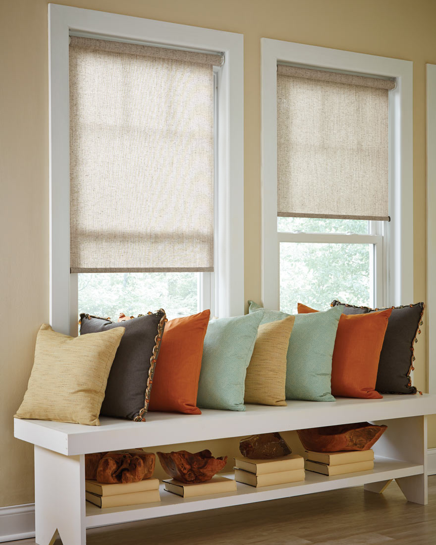 two light beige Genesis® Roller Shades with a bench that has several brightly colored Interior Masterpieces® Custom Pillows