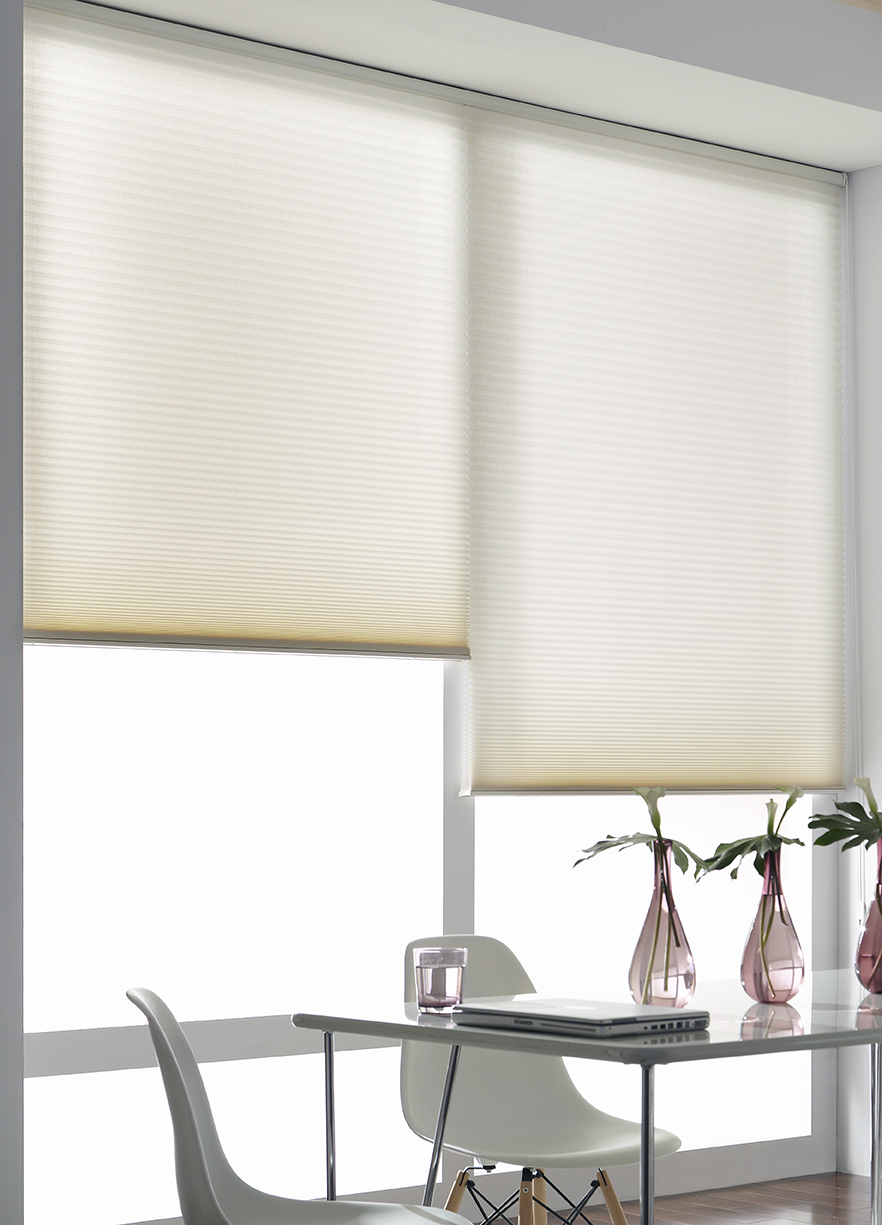 Two cream cellular shades hang on one headrail in a large window of a bright, light modern dining and work space area.