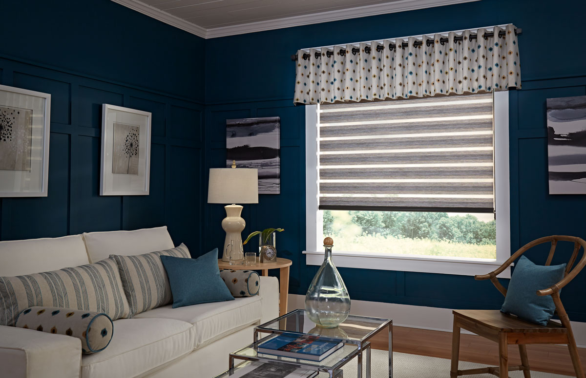 light brown Allure® transitional shade with custom Interior Masterpieces® valance hanging on a black custom rod with grommets and finials in a blue room with a cream colored couch that has accent pillows on it