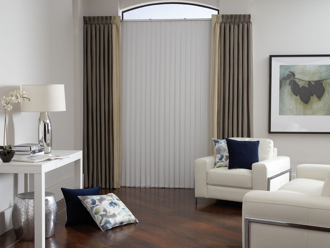 White Sheer Vision® Vertical Blinds with brown and tan custom draperies and pillows from our Interior Masterpieces® collection in a room with white chairs and a table