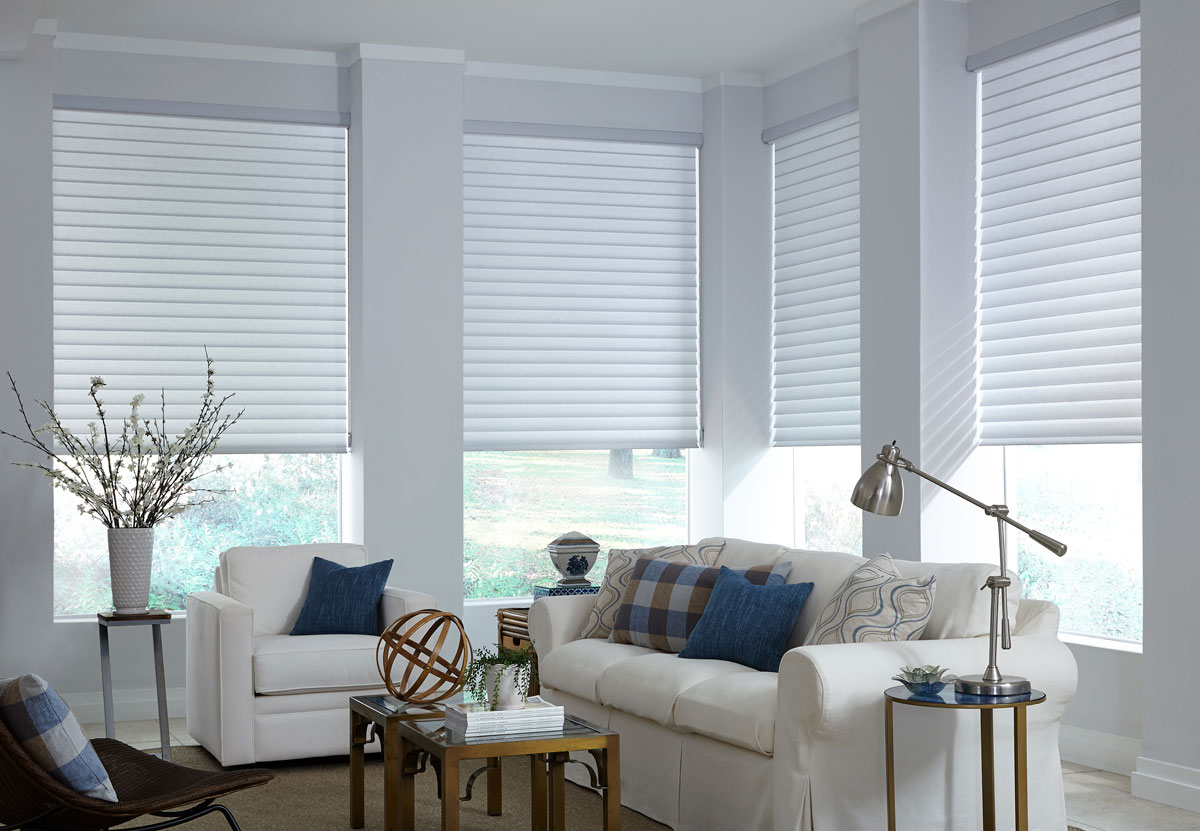 Several white Tenera® Sheer Shades half open in a room with white walls and a white couch with a white chair that have blue and blue plaid Interior Masterpieces® custom pillows