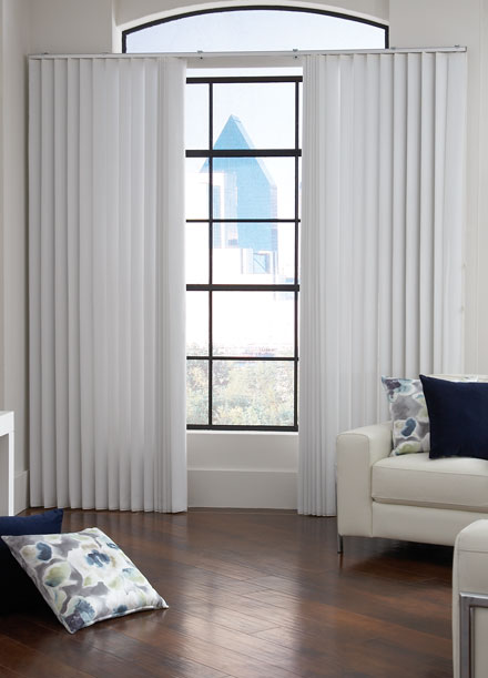 White sheer fabric wrapped vertical blinds hang in a window of a room with dark wood floors, modern white leather chairs with metal accents and custom throw pillows in navy and floral.