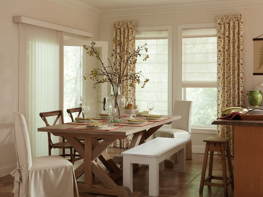 white Discoveries® Vertical Blinds on one large patio door with custom Genesis® shades with Interior Masterpieces® draperies on the other wall in a kitchen with a brown table and white covered chairs